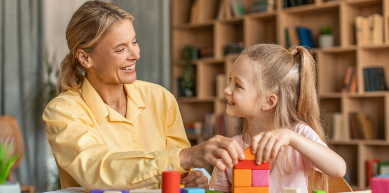 BCBA smiling and helping a young girl build colorful blocks at a table during ABA therapy session.