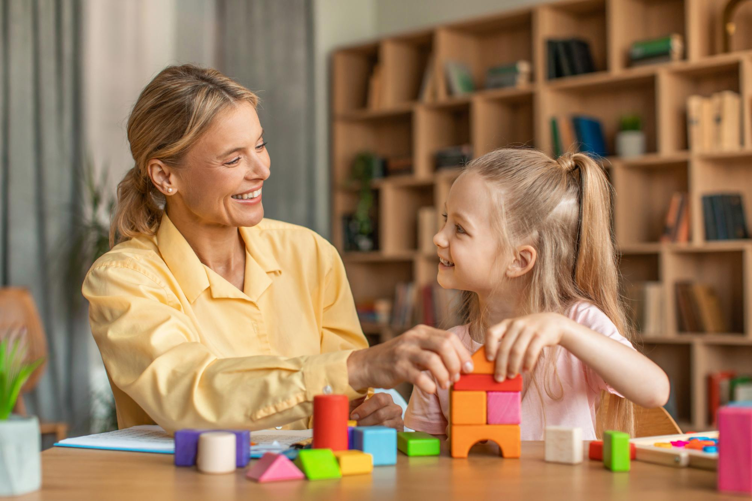 BCBA smiling and helping a young girl build colorful blocks at a table during ABA therapy session.
