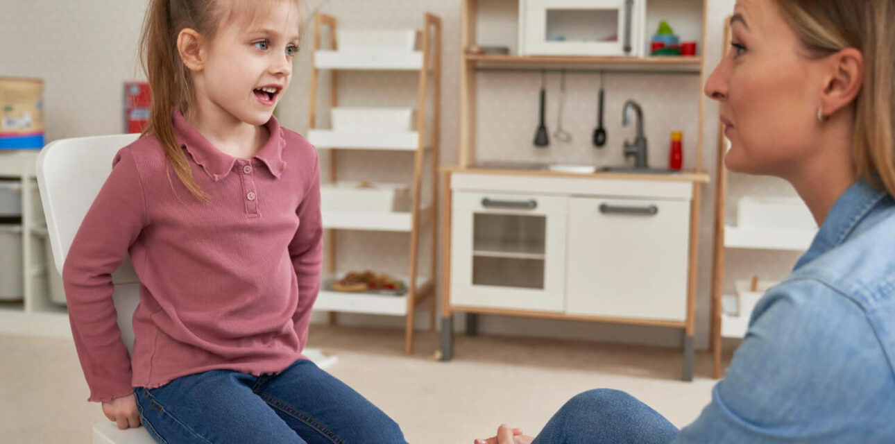 Young girl talking with an ABA therapist in a playroom with a toy kitchen.