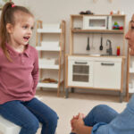 Young girl talking with an ABA therapist in a playroom with a toy kitchen.