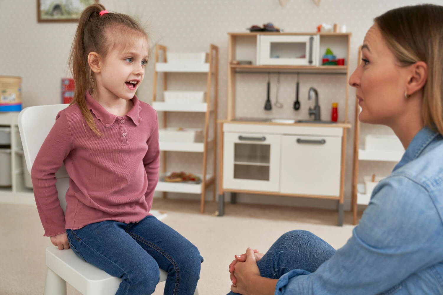 Young girl talking with an ABA therapist in a playroom with a toy kitchen.