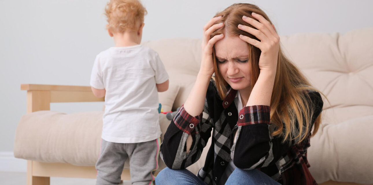 Parent sitting on the floor looking stressed while a young child stands nearby.