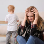 Parent sitting on the floor looking stressed while a young child stands nearby.