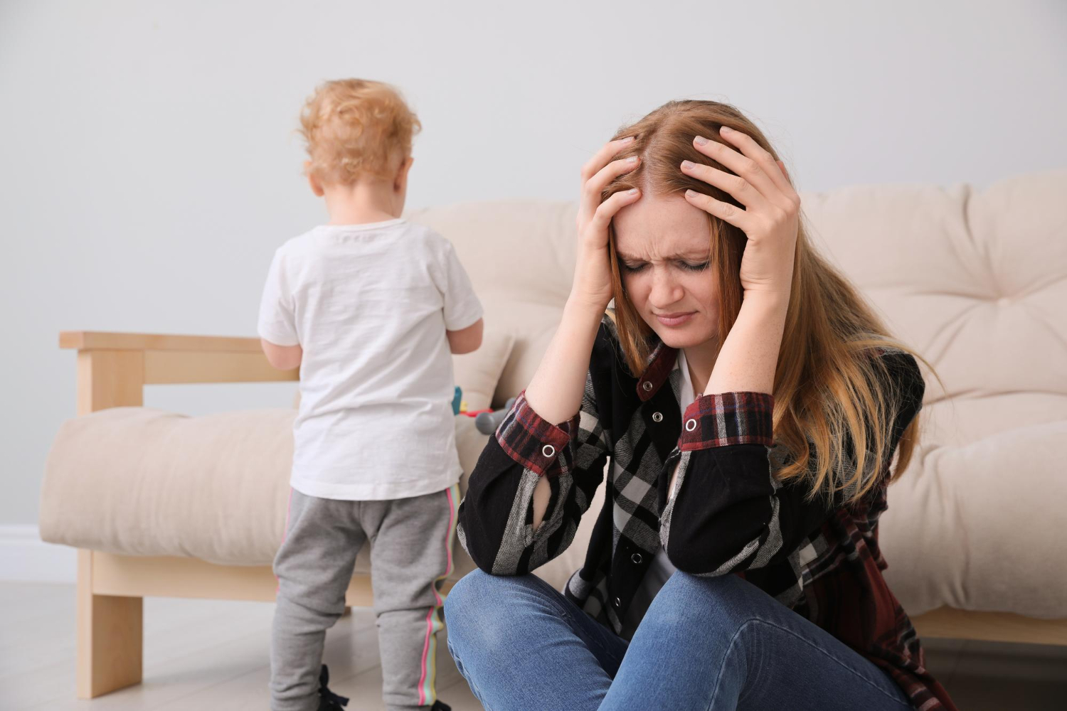 Parent sitting on the floor looking stressed while a young child stands nearby.