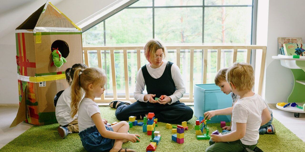 Teacher and four young children sitting on a rug, building colorful block towers in a bright classroom.