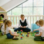 Teacher and four young children sitting on a rug, building colorful block towers in a bright classroom.