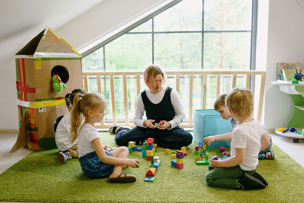 Teacher and four young children sitting on a rug, building colorful block towers in a bright classroom.
