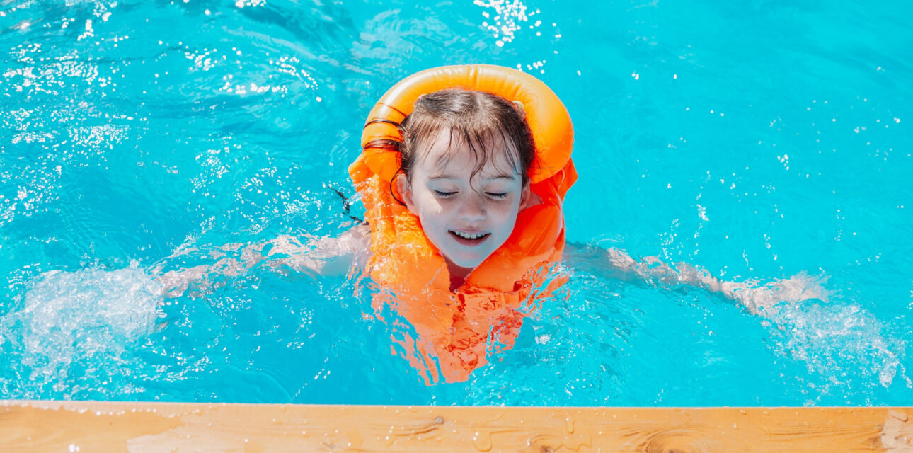 Child with autism wearing an orange life jacket swimming in a bright blue pool near the pool edge.