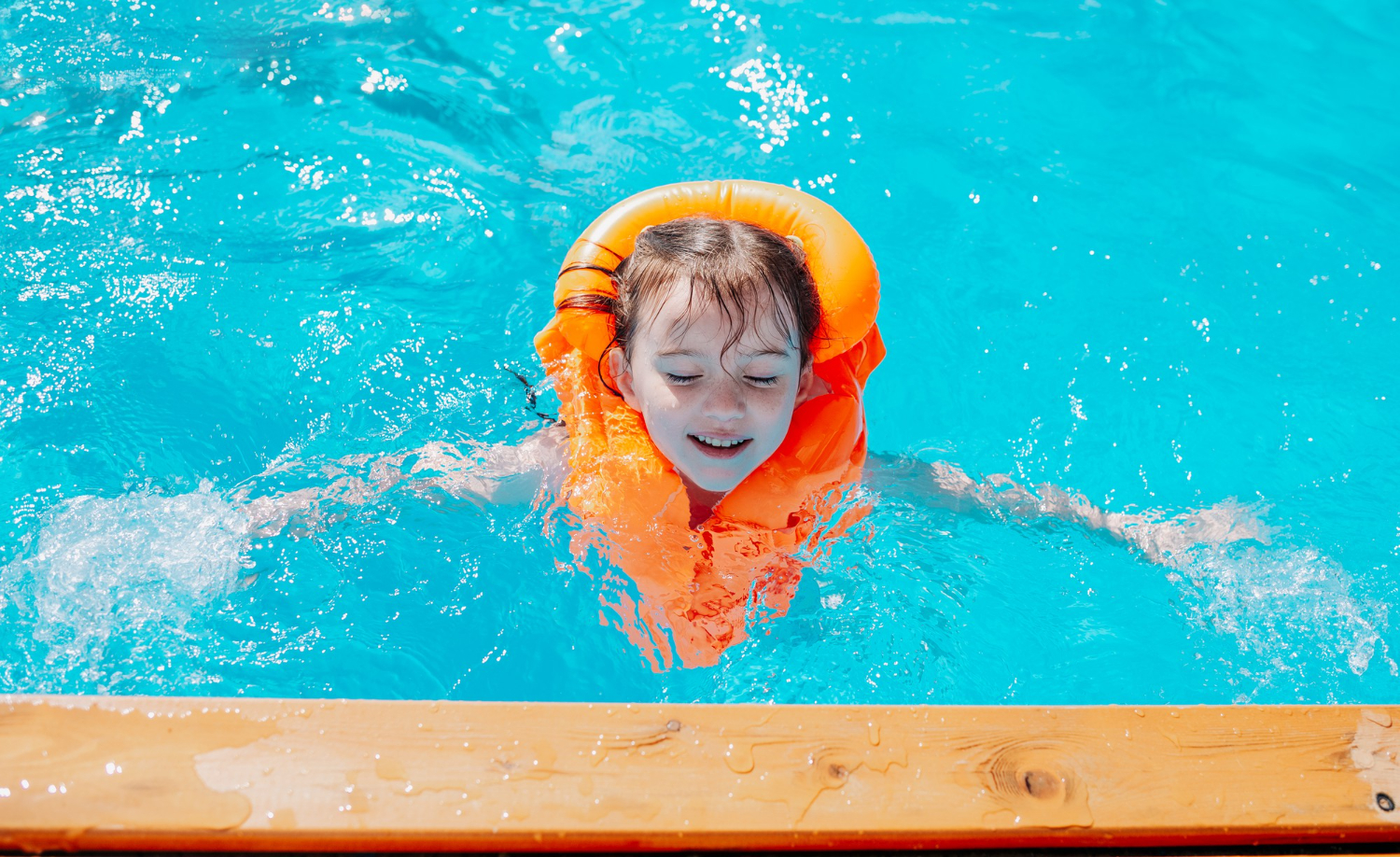 Child with autism wearing an orange life jacket swimming in a bright blue pool near the pool edge.