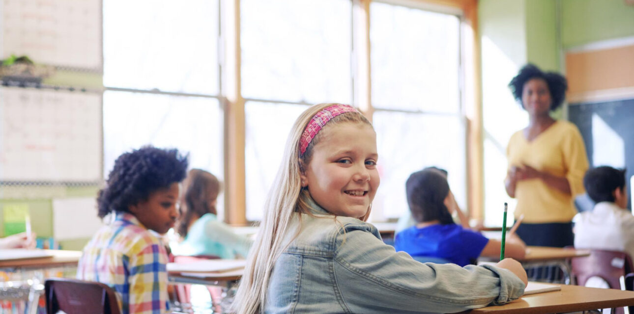 Autistic student smiling at the camera while sitting at a desk in a classroom.
