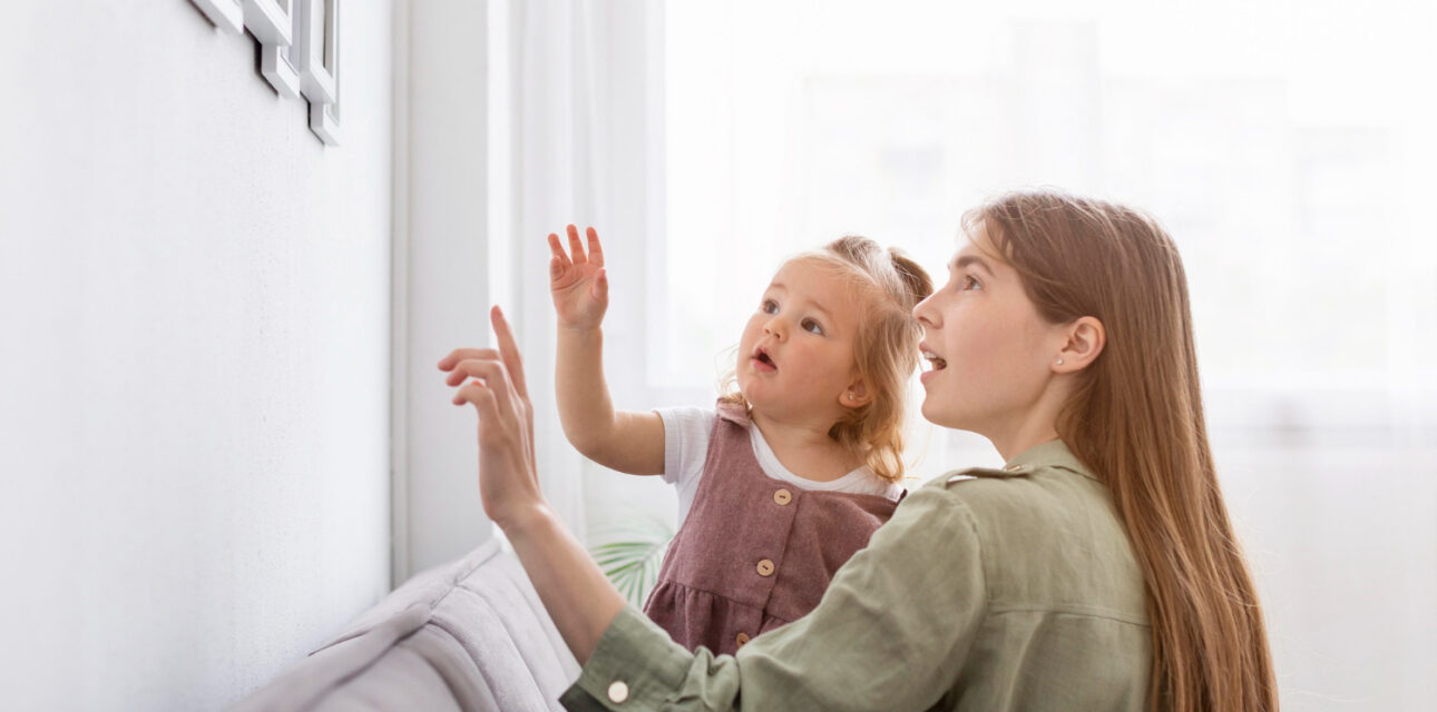 A BCBA holding a toddler with autism as they point and look at framed photos on a wall.