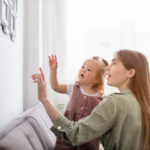 A BCBA holding a toddler with autism as they point and look at framed photos on a wall.