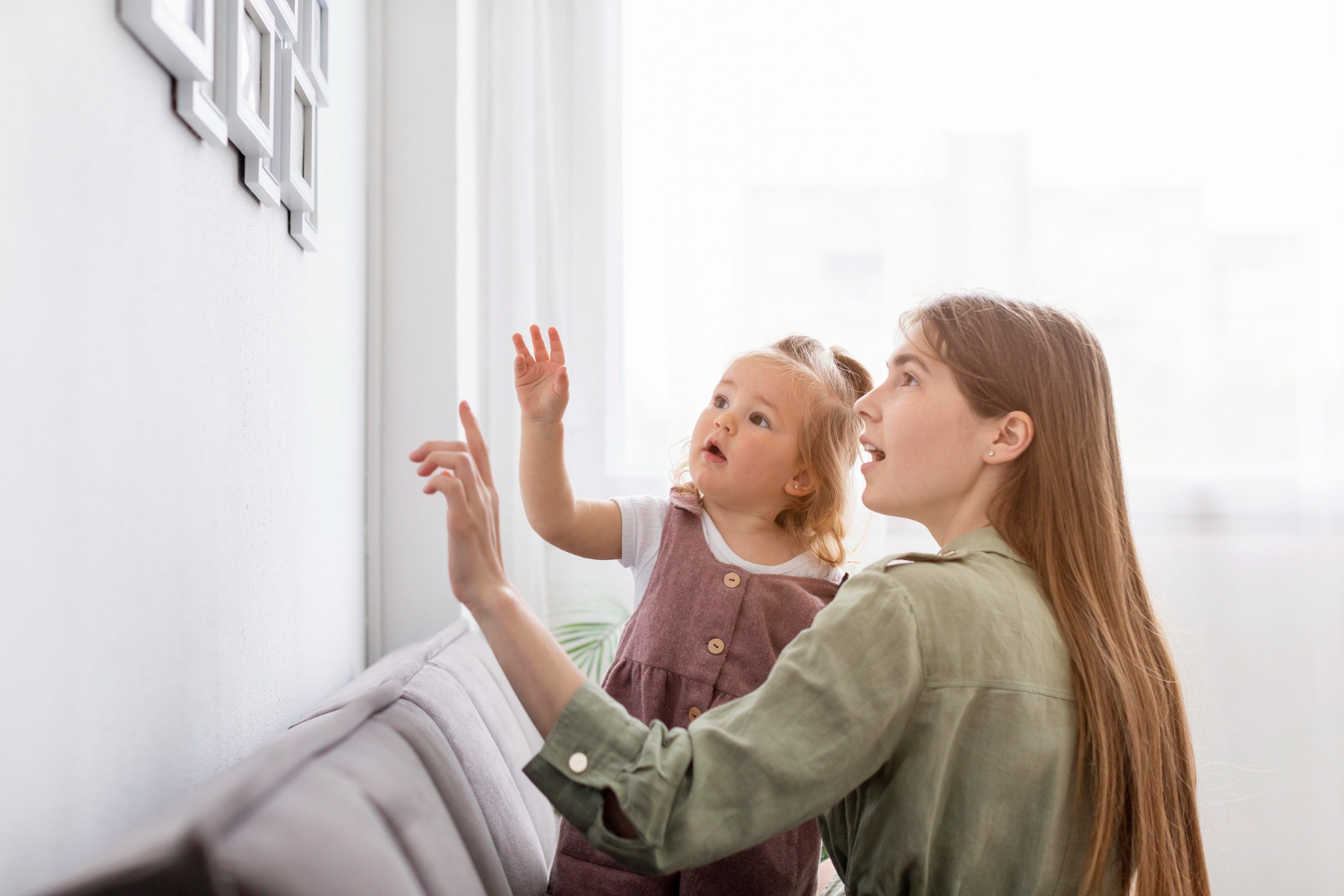 A BCBA holding a toddler with autism as they point and look at framed photos on a wall.