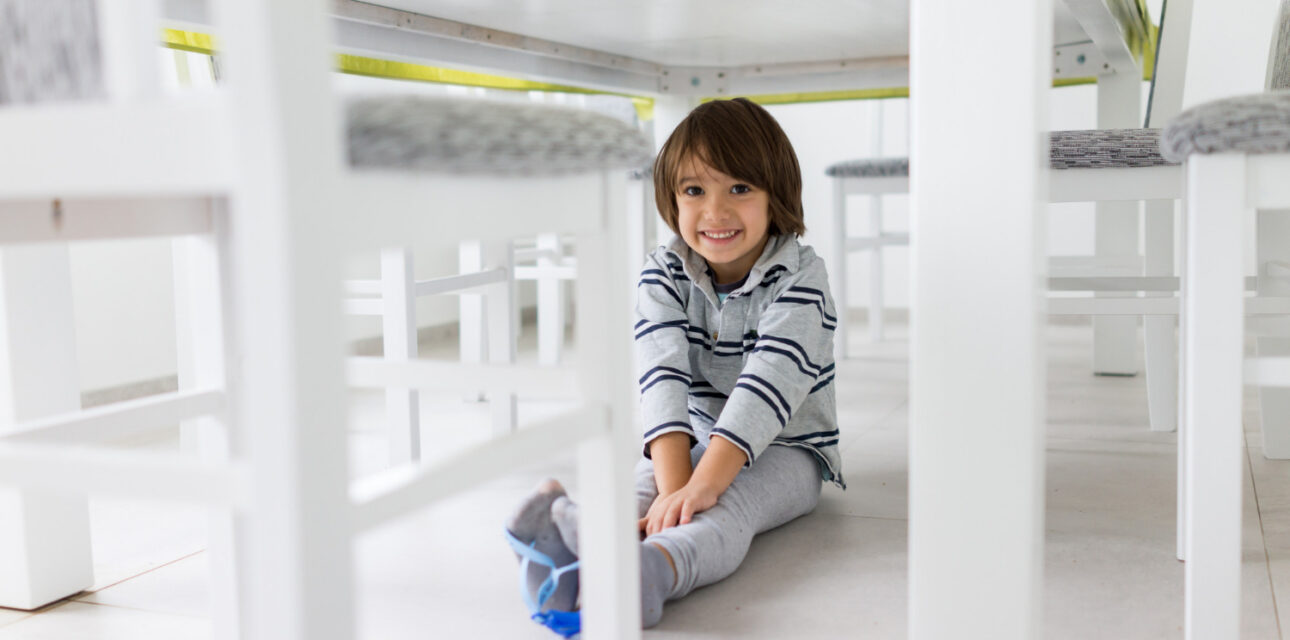 Smiling young child sitting on the floor under a white dining table, surrounded by chairs.