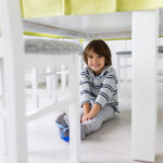 Smiling young child sitting on the floor under a white dining table, surrounded by chairs.