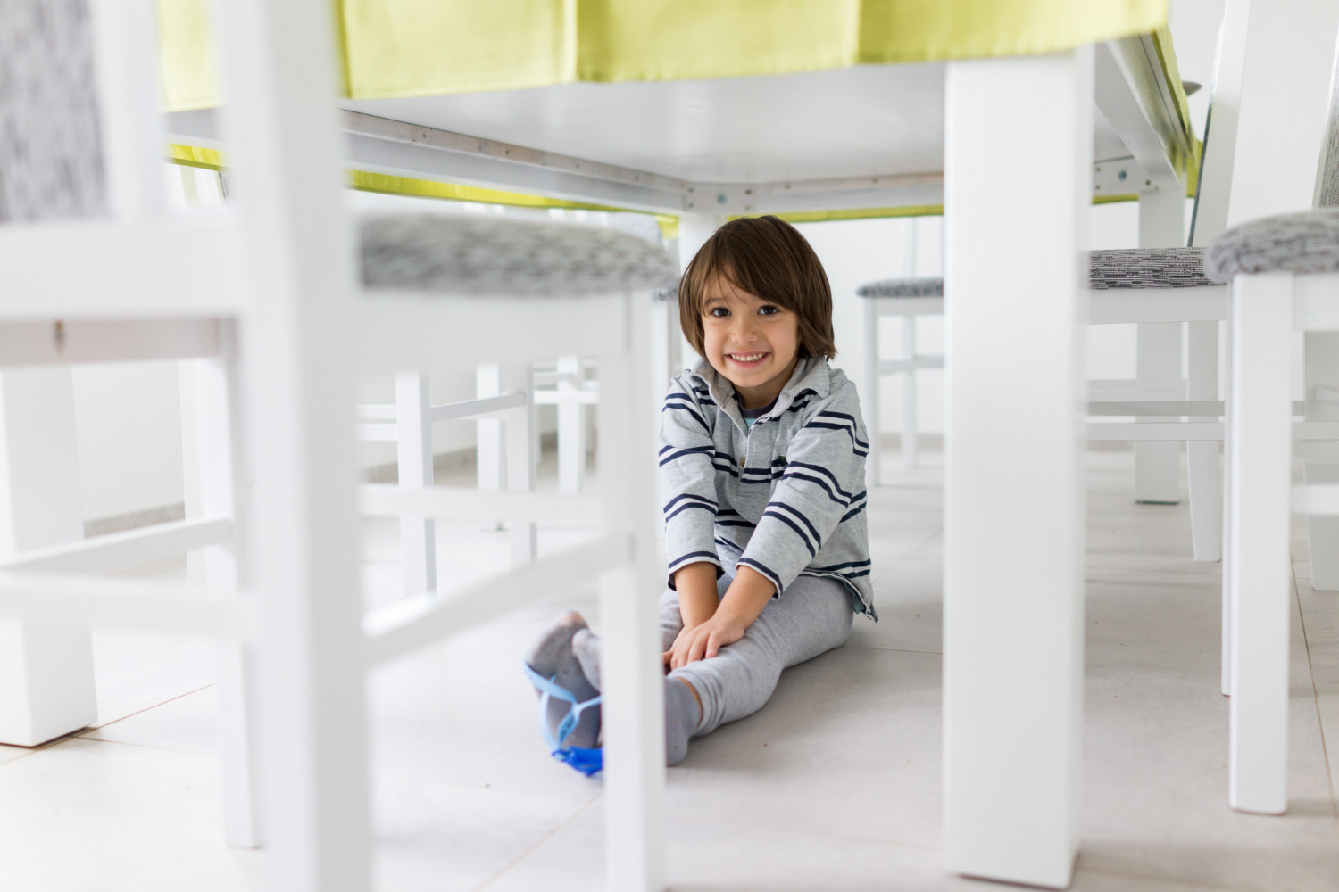 Smiling young child sitting on the floor under a white dining table, surrounded by chairs.