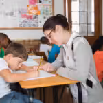 Teacher providing one-on-one school-based ABA support to a student with autism during a classroom writing activity.