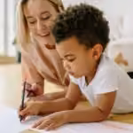 Child drawing with colored pencils alongside a supportive adult during a play-based ABA therapy activity.