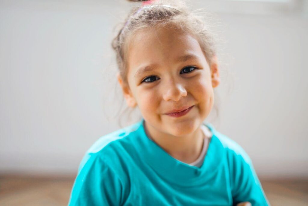 Smiling young girl in a turquoise shirt looking at the camera indoors.