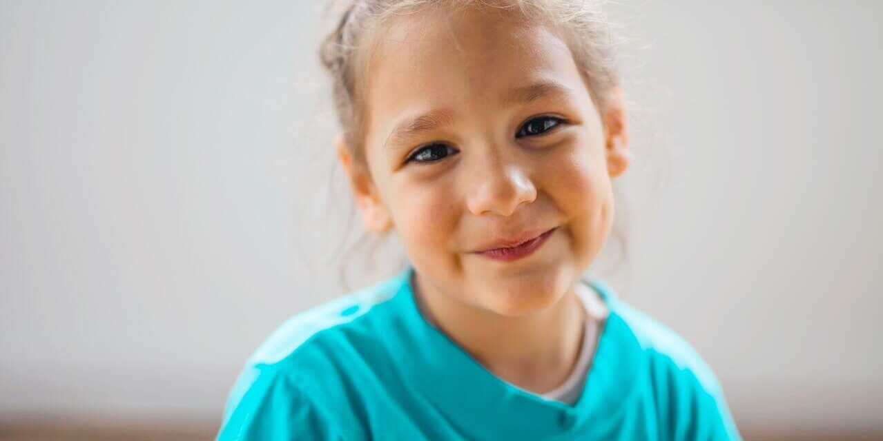 Smiling young girl in a turquoise shirt looking at the camera indoors.