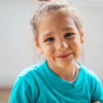 Smiling young girl in a turquoise shirt looking at the camera indoors.