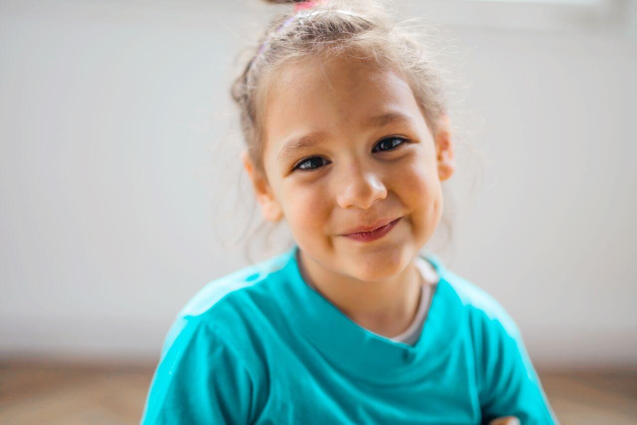 Smiling young girl in a turquoise shirt looking at the camera indoors.