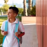 School-aged autistic boy with a backpack walking outside a school building.